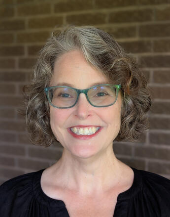 A woman with a welcoming smile, glasses and short, gray/brown, curly hair.